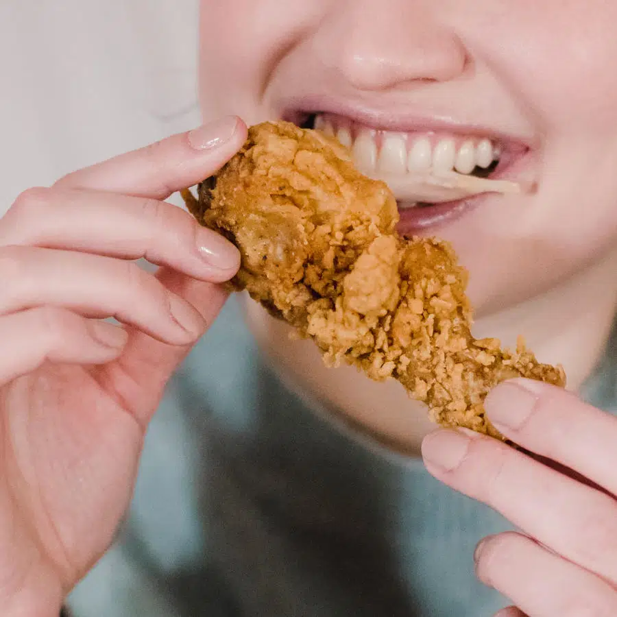 Eine junge Frau hält ein frittiertes Stück Hühnchen in den Händen und beißt gerade davon ab.