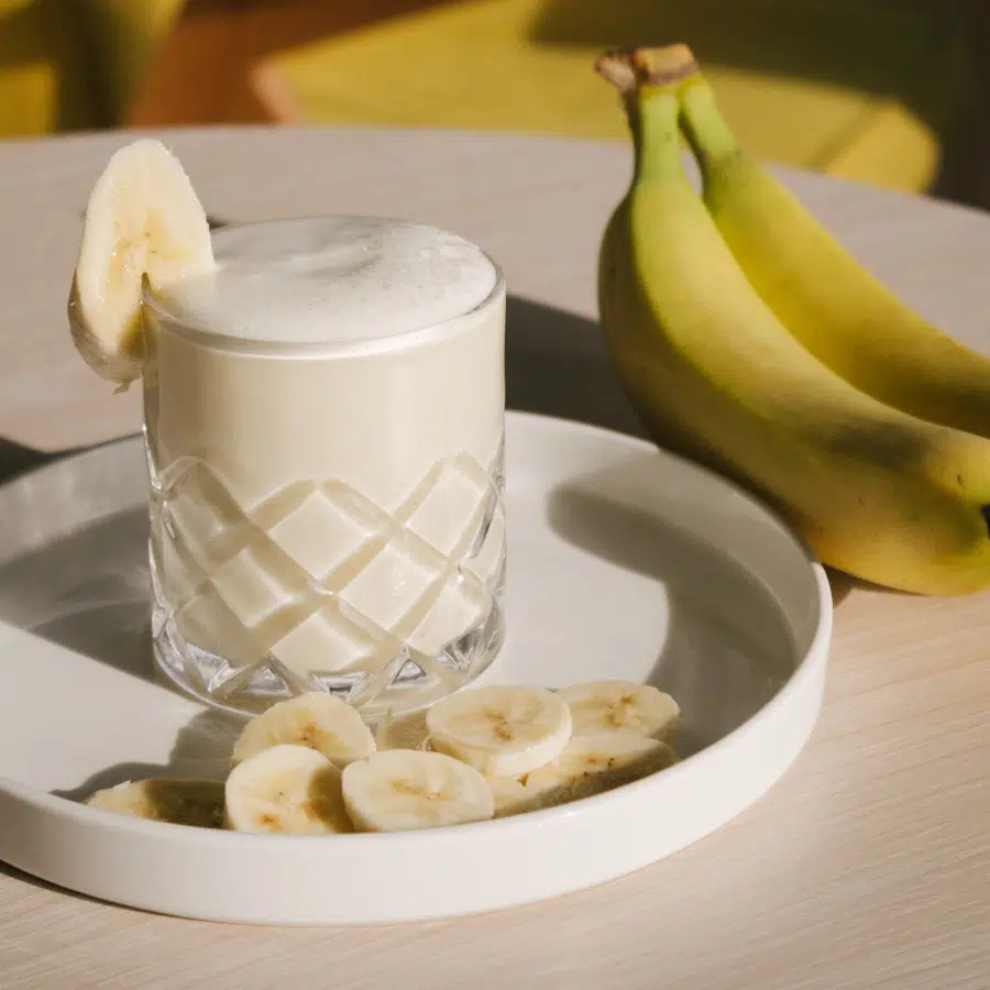 Ein Glas mit selbstgemachter Bananenmilch steht auf einem runden weißen Teller. Daneben liegen geschnittene Bananenscheiben und im Hintergrund zwei ganze Bananen.