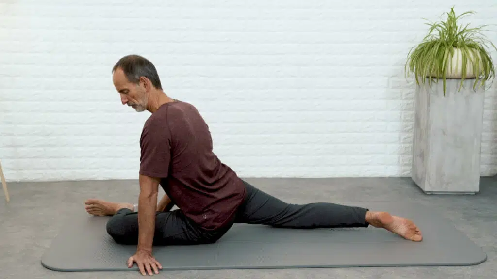 Roland Liebscher-Bracht sitzt auf seiner linken Gesäßhälfte und hat das linke Bein nach vorne angewinkelt. Im Knie ist ein rechter Winkel. Das rechte Bein ist lang nach hinten ausgestreckt und berührt mit dem Fußspann den Boden. Er stützt sich mit der linken Hand vom Boden ab. Der Oberkörper ist aufgerichtet.