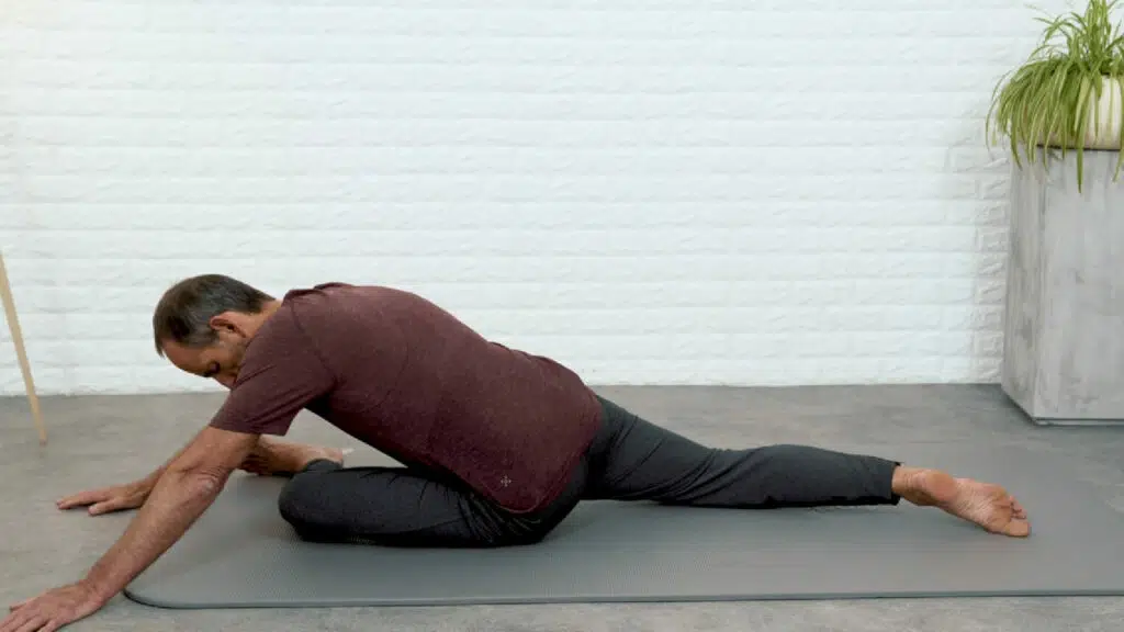 Roland Liebscher-Bracht sitzt auf seiner linken Gesäßhälte und hat das linke Bein nach vorne angewinkelt. Im Knie ist ein rechter Winkel. Das rechte Bein ist lang nach hinten ausgestreckt und berührt mit dem Fußspann den Boden. Er stützt sich mit der linken Hand vom Boden ab. Der Oberkörper ist nach vorne gestreckt.