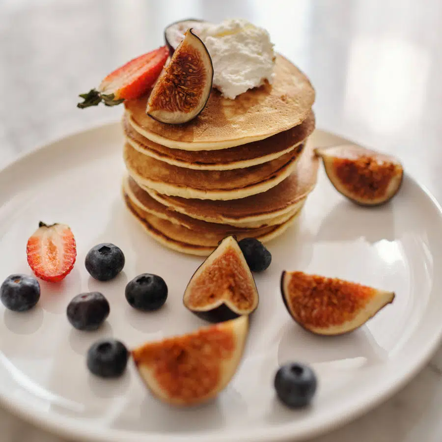 Ein Stapel gesunder Pfannkuchen liegt auf einem Teller. Drumherum sind Blaubeeren, eine aufgeschnittene Erdbeere und frische Feigen verteilt. Auf dem Stapel Pfannkuchen ist außerdem ein Klecks Sojacreme und eine halbe Erdbeere und ein Stück Feige.