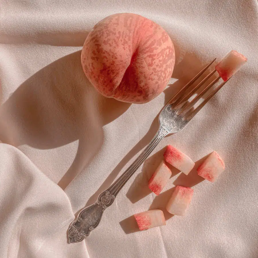 Ein Pfirsich liegt auf einem lachsfarbenen Tuch. Unter ihm liegt eine Gabel, auf deren Zinken ein Stück Pfirsich steckt. Darunter liegen weitere Pfirsichstücke.