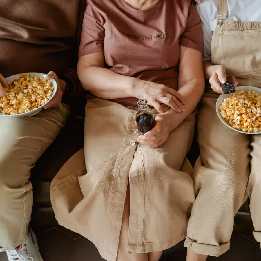 Drei Personen sitzen eng beieinander auf einer Couch. Die Frau in der Mitte hält eine Colaflasche in der Hand, während die beiden Personen links und rechts eine Schale Popcorn halten.