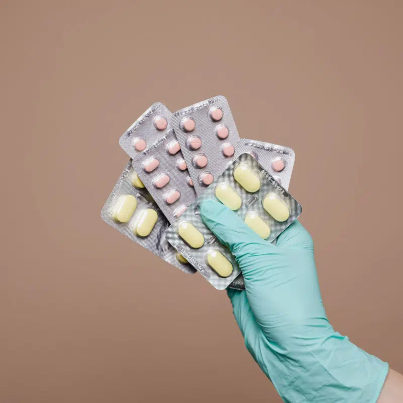 Eine Hand in einem türkisen Latexhandschuh hält mehrere Tablettenpackungen in die Luft. Die Tabletten haben verschiedene Formen und sind gelb und rosa. Der Hintergrund ist braun.
