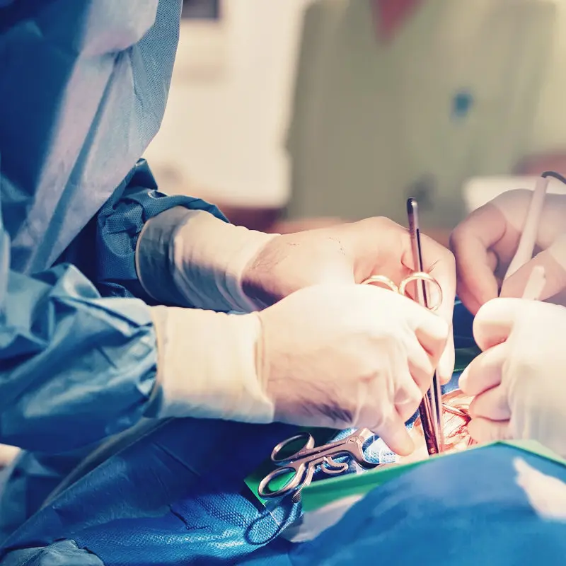 Chirurg:innen operieren einen Patienten an der Schulter. Sie tragen weiße Latexhandschuhe und halten diverse metallische Instrumente in den Händen.