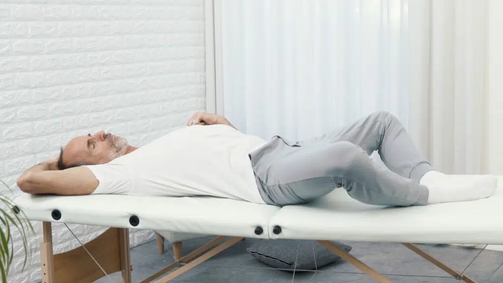 Roland Liebscher-Bracht liegt auf einer therapeutischen Liege, die Füße aneinander und die Knie fallen zu den Seiten. Mit einer Hand an seinem Kopf zieht er seinen Kopf zu Schulter, um den seitlichen Nacken zu dehnen.