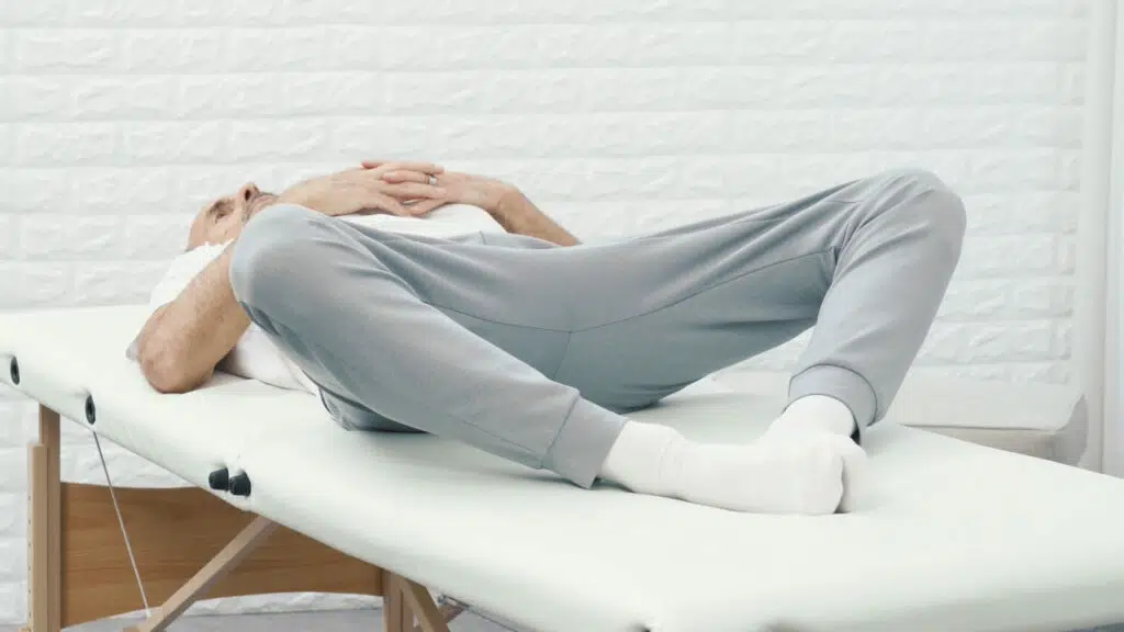 Roland Liebscher-Bracht liegt mit dem Rücken auf einer therapeutischen Liege. Seine Fußsohlen liegen aneinander, sodass seine Knie zu den Seiten fallen.