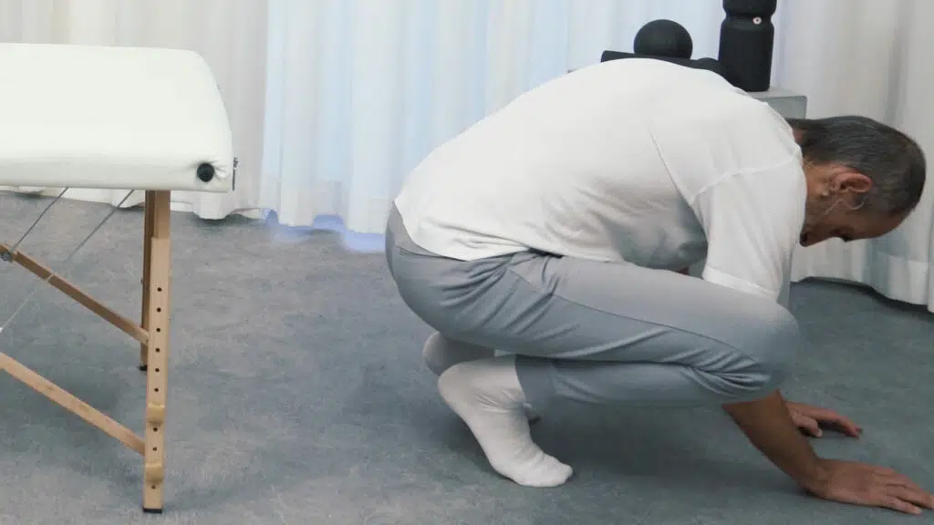 Roland Liebscher-Bracht sitzt am Ende einer therapeutischen Liege und geht in die Hocke.