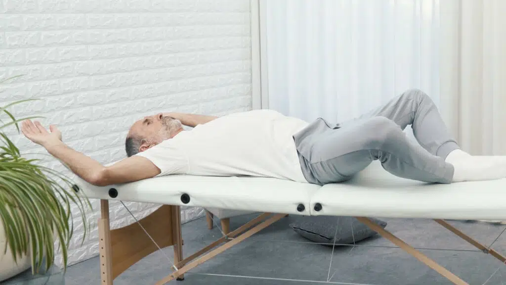 Roland Liebscher-Bracht liegt auf einer therapeutischen Liege, seine Fußsohlen liegen aneinander, seine Knie fallen zu den Seiten und seine Arme streckt er nach oben und hinten.
