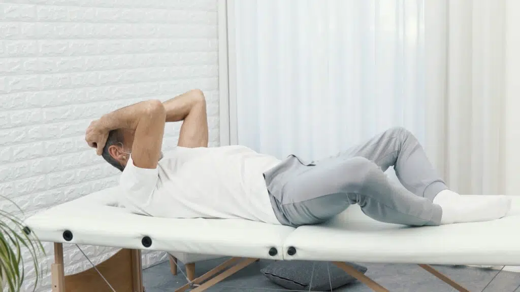 Roland Liebscher-Bracht liegt auf einer therapeutischen Liege, seine Fußsohlen liegen aneinander, seine Knie fallen zu den Seiten und mit den Händen am Hinterkopf zieht er sein Kinn zur Brust.