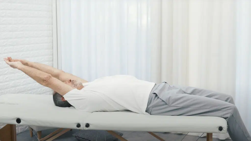 Roland Liebscher-Bracht liegt auf einer therapeutischen Liege. Seine Beine lässt er ab der Kniekehle nach unten baumeln. Seine Arme streckt er lang nach hinten aus.