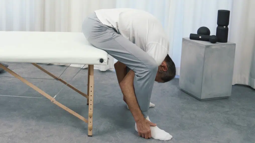 Roland Liebscher-Bracht sitzt am Ende einer therapeutischen Liege und beugt sich mit dem Oberkörper nach unten, sodass er seine Füße greifen kann.