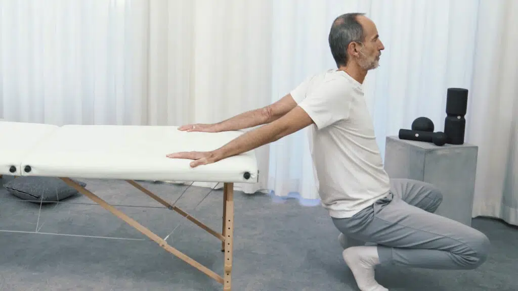 Roland Liebscher-Bracht befindet sich am Ende einer therapeutischen Liege, er geht in die Knie, während seine Arme ausgestreckt hinten auf der Liegefläche bleiben.