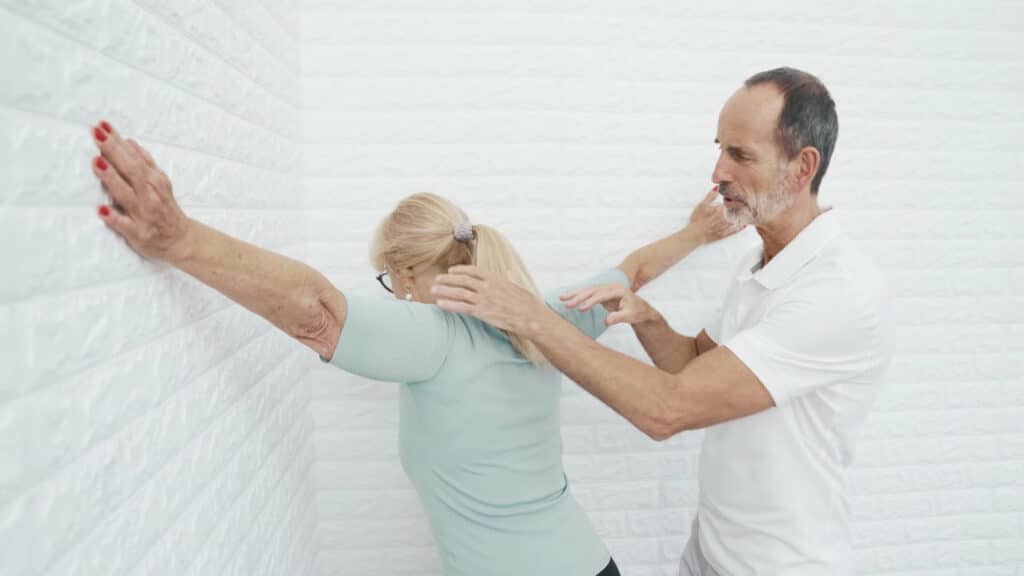 Die Patientin steht in einer Zimmerecke. Sie hat die Arme zu den Seiten ausgebreitet und gestreckt. Die Handflächen berühren die Wand. Ihr Brustbein lehnt sie langsam in die Ecke hinein.