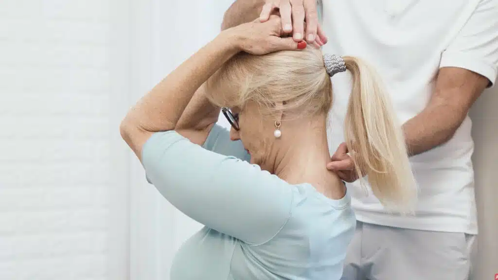 Die Patientin umgreift mit beiden Händen ihren Hinterkopf. Sie hält den Oberkörper sehr aufrecht und zieht mit beiden Händen den Kopf langsam nach vorne und unten, sodass sie ein Doppelkinn macht.