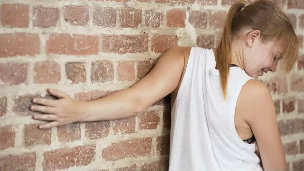 Eine junge Frau steht an einer gemauerten Wand. Ihren rechten Arm hält sie lang ausgestreckt gegen die Wand, während sie ihren Oberkörper nach außen aufdreht.