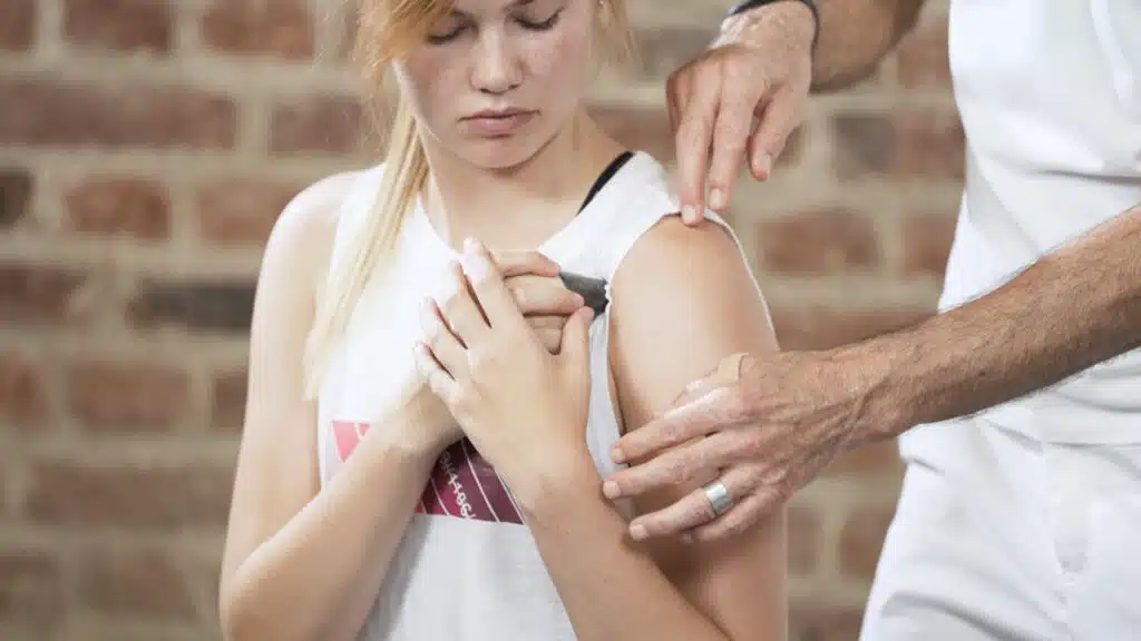 Eine junge Frau hält sich den Drücker an einen Punkt der Schulter-Vorderseite. Roland Liebscher-Bracht steht neben ihr und korrigiert ihre Position.