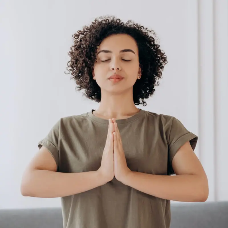 Eine Person hat die Handflächen vor dem Brustkorb aufeinander gelegt und die Augen geschlossen. Sie trägt ein grau-braunes T-Shirt und hat kurze schwarze Locken.