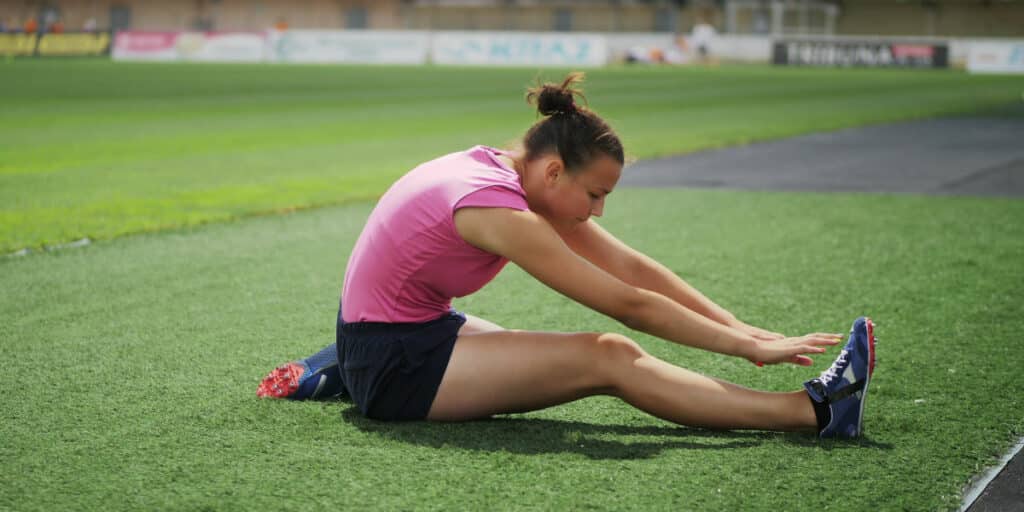 Leichtathletin auf Rasen in Stadion macht Dehnübung, rechtes Bein ausgestreckt, linkes Bein angewinkelt, greift mit beiden Händen zum rechten Fuß