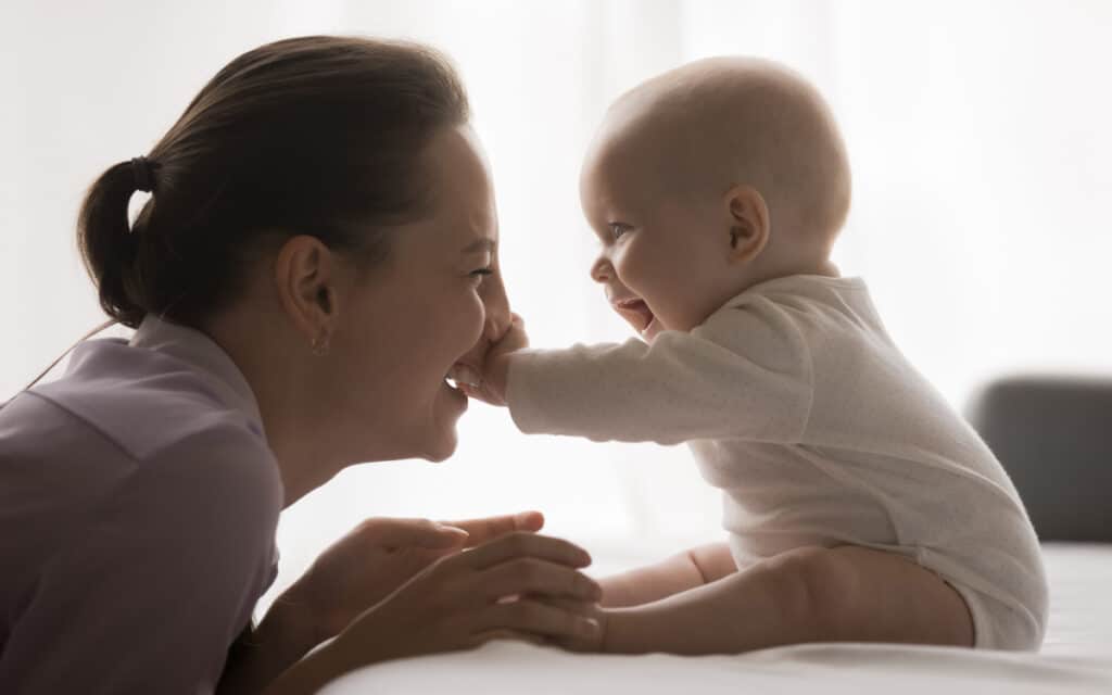 Baby sitzt auf Tisch, Frau greift Füßchen, Baby greift Nase der Frau, beide lachen