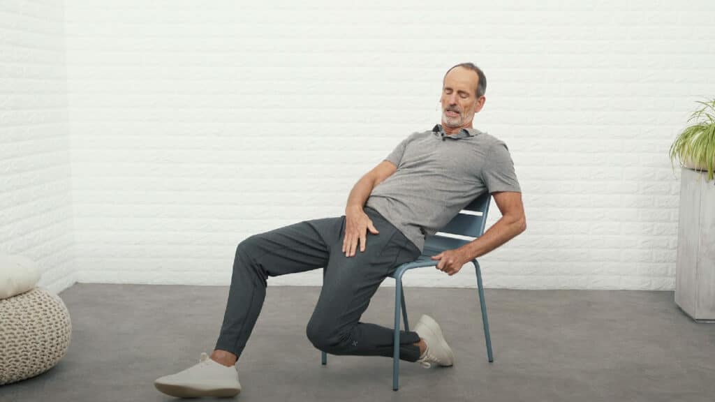 Roland Liebscher-Bracht sitzt sehr weit vorne auf einem Stuhl und hat das linke Bein so angewinkelt, dass es unter dem Stuhl liegt. Der linke Fuß liegt mit dem Fußrücken am Boden. Der rechte Fuß ist vor dem Stuhl aufgestellt.