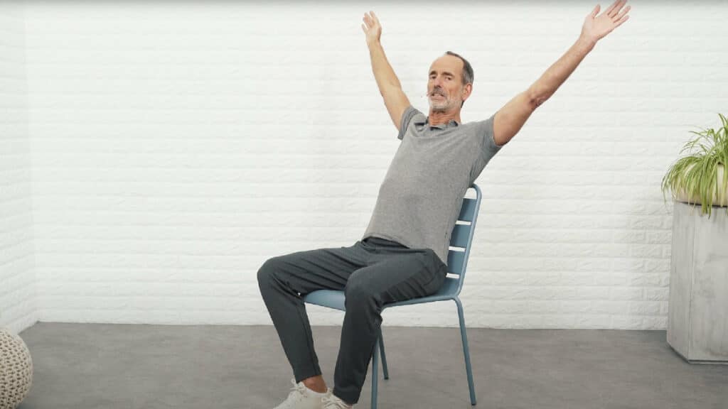 Roland Liebscher-Bracht hat beide Arme erhoben und streckt sie v-förmig geöffnet nach oben und hinten. Dabei lehnt er sich gegen die Stuhllehne.