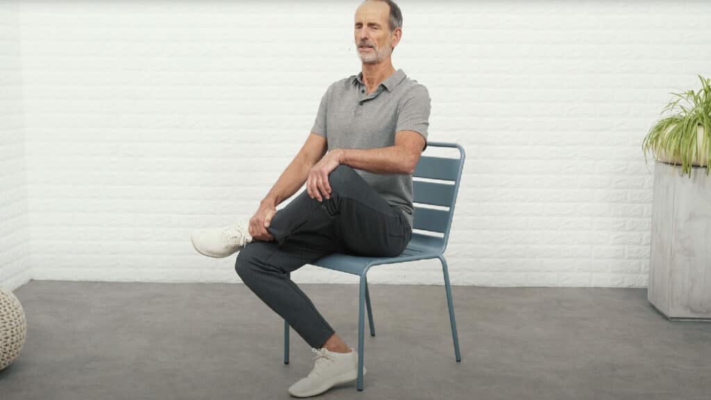 Roland Liebscher-Bracht sitzt auf einem Stuhl und hat das Sprunggelenk des linken Fußes auf dem rechten Knie abgelegt. Das linke Knie schwebt in der Luft, er hält es mit der linken Hand fest. Die rechte Hand ist am Sprunggelenk. Der Rücken ist sehr gerade und er lehnt sich leicht nach vorne.