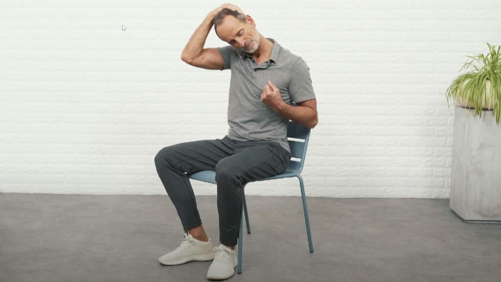 Roland Liebscher-Bracht sitzt auf einem Stuhl und hat den linken Arm so angewinkelt, dass der Ellbogen eng an seinem Körper ist und die Faust etwa auf Brusthöhe. Die rechte Hand greift über den Kopf und zieht den Kopf nach recht unten. Dabei ist das Gesicht zur linken Faust ausgerichtet.