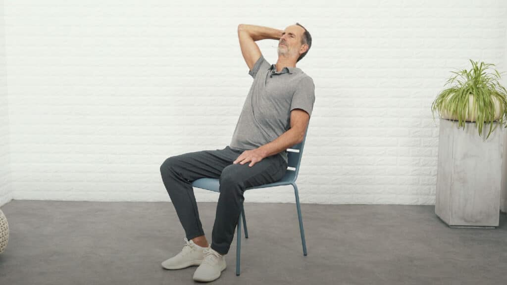 Roland Liebscher-Bracht sitzt auf einem Stuhl und lehnt sich nach hinten gegen die Stuhllehne. Er hat sein Kinn dicht an den Hals gezogen und schiebt seinen Hinterkopf nach hinten in die rechte Hand hinein.