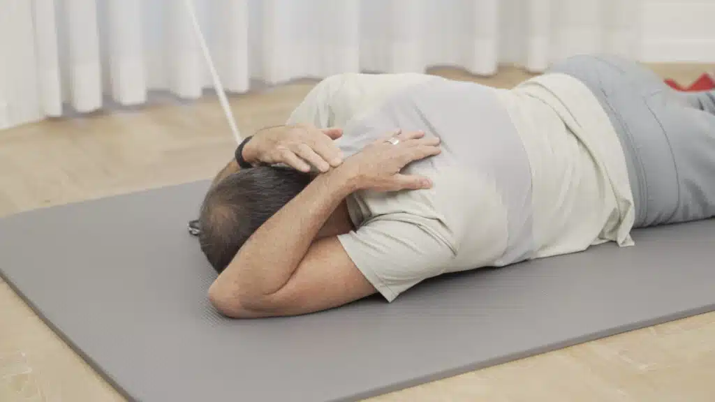 Roland Liebscher-Bracht liegt auf dem Bauch auf einer Matte und hat den linken Arm nach vorne gelegt und gebeugt. Seine linke Hand berührt sein Schulterblatt, der gebeugte Ellbogen berührt die Matte neben seinem Kopf. Mit der rechten Hand drückt er seine linke Hand näher ans Schulterblatt