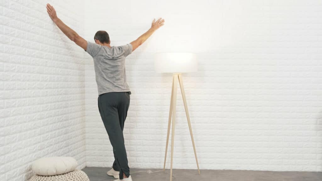 Roland Liebscher-Bracht steht in einer Zimmerecke und hat die Arme etwas höher als Schulterhöhe zu den Seiten ausgebreitet. Die Hände berühren die Wände. Er macht einen Ausfallschritt und lehnt sein Brustbein in die Ecke hinein.