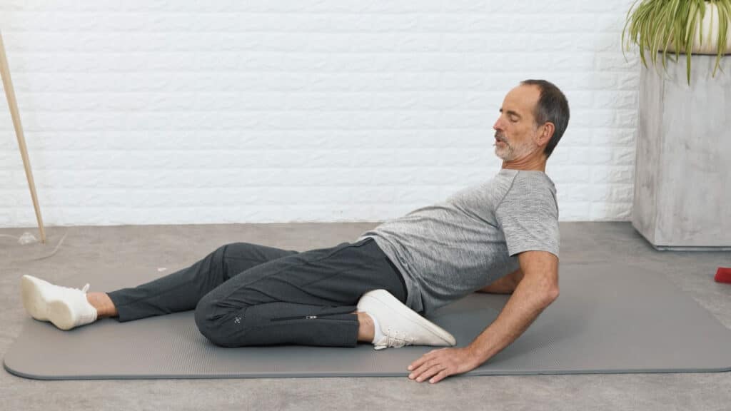 Roland Liebscher-Bracht sitzt auf dem Boden und hat das linke Bein angewinkelt. Seine linke Ferse liegt etwa am Gesäß links. Er stützt sich hinten mit den Armen auf der Matte ab und lehnt sich zurück.