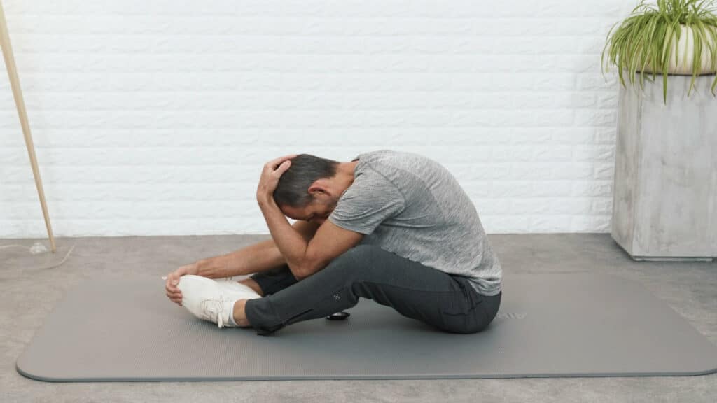 Roland Liebscher-Bracht sitzt auf dem Boden und hat die Beine vor sich zu den Seiten angewinkelt. Die Fußsohlen berühren sich. Eine Hand hält die Füße fest, die andere Hand zieht den Kopf nach unten in eine Vorbeuge.