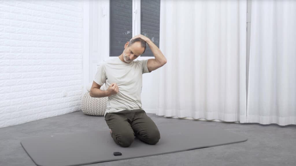 Roland Liebscher-Bracht kniet auf einer Matte und hat den rechten Arm so angewinkelt, dass der Ellenbogen an seiner Hüfte ist und die Faust auf Brusthöhe. Die rechte Schulter zieht er runter, während die linke Hand den Kopf umgreift und den Kopf nach links zieht.