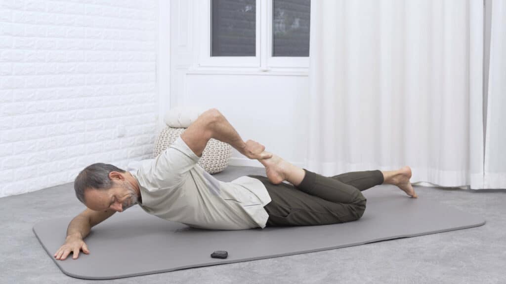 Roland Liebscher-Bracht liegt auf dem Bauch und hat sein linkes Bein angewinkelt. Er hält den Fuß des Beines fest und zieht ihn in Richtung Gesäß heran.