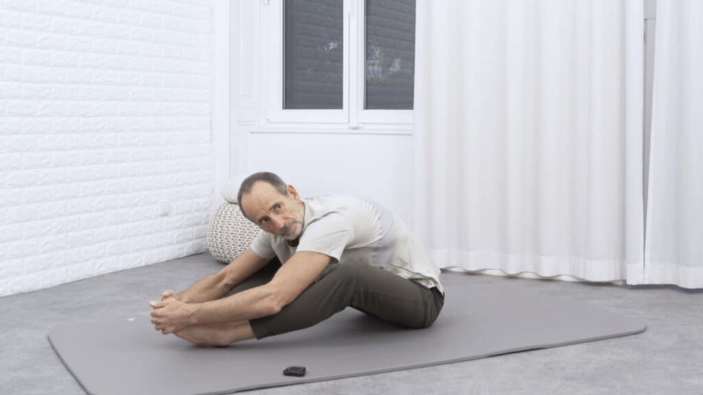 Roland Liebscher-Bracht sitzt auf dem Boden und hat beide Beine vor sich angewinkelt, sodass die Fußsohlen zueinander zeigen. Die Beine sind etwa einen Meter von ihm entfernt. Mit beiden Händen greift er die Füße und lehnt sich nach vorne.