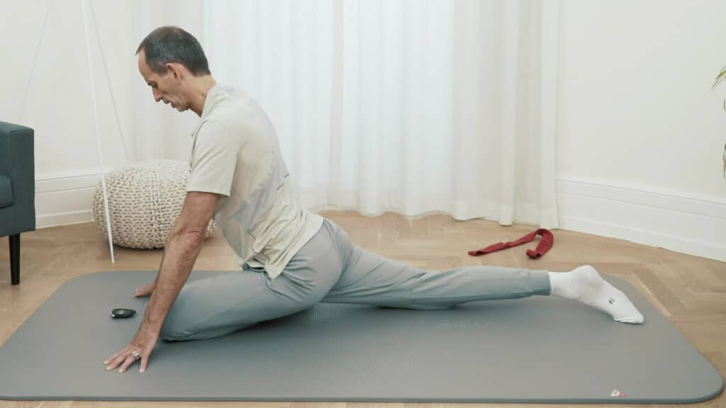 Roland Liebscher-Bracht hat das linke Bein nach vorne angewinkelt und das rechte Bein nach hinten ausgestreckt. Er ist in der Yogaposition "Taube". Mit den Fingerspitzen stützt er den Oberkörper ab.