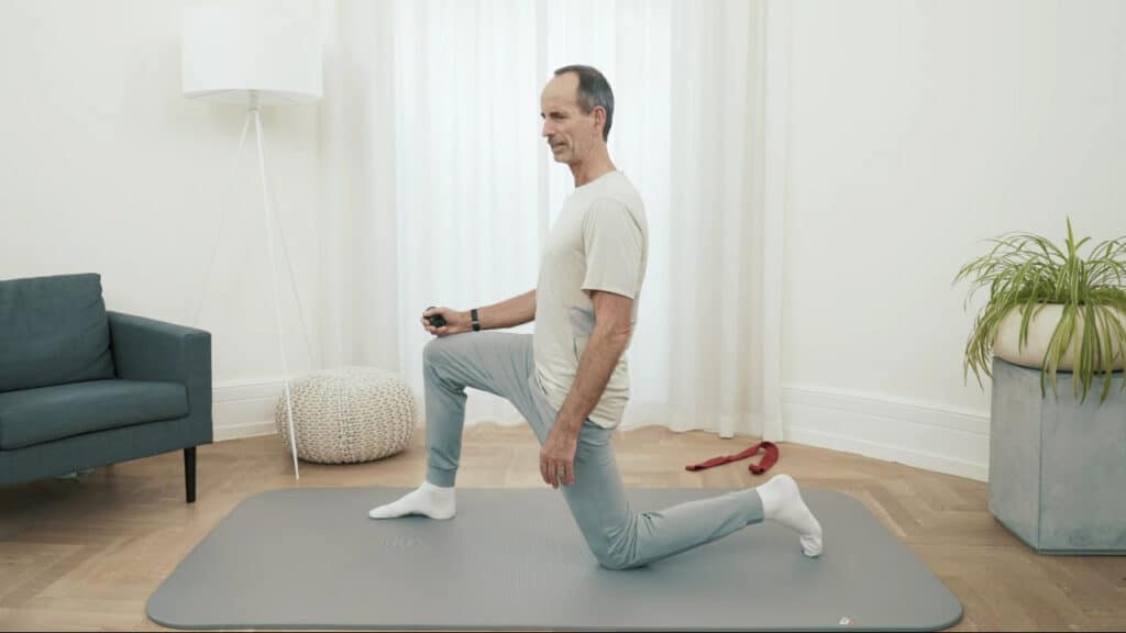 Roland Liebscher-Bracht hat das linke Knie auf einer Matte abgestellt und mit dem rechten Fuß einen Ausfallschritt gemacht. Der Oberkörper ist aufgerichtet.