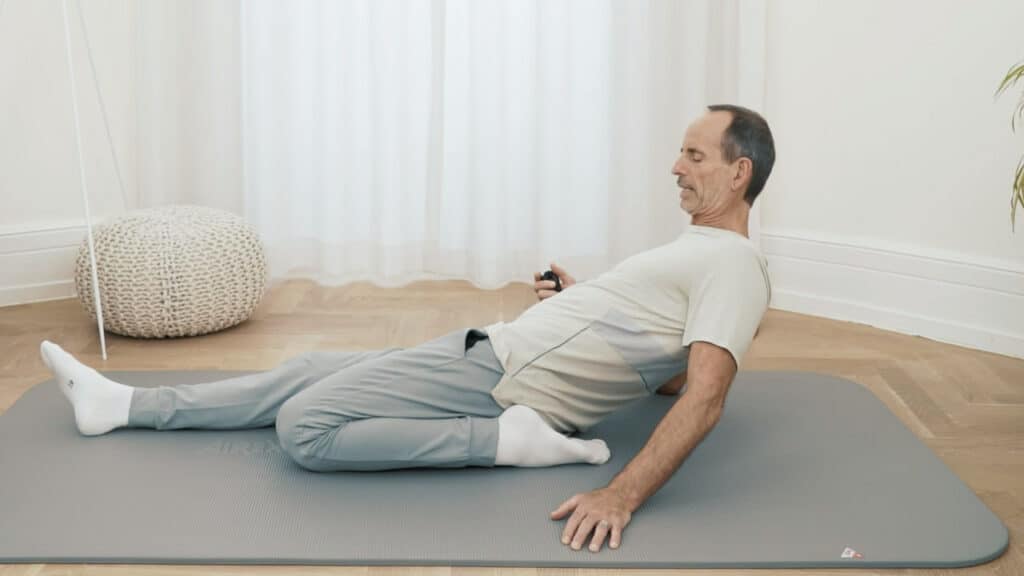 Roland Liebscher-Bracht sitzt auf dem Boden und hat das linke Bein nach hinten angewinkelt. Das rechte Bein ist nach vorne ausgestreckt. Sein linker Fuß ist nah am Gesäß und er lehnt sich nach hinten zurück.