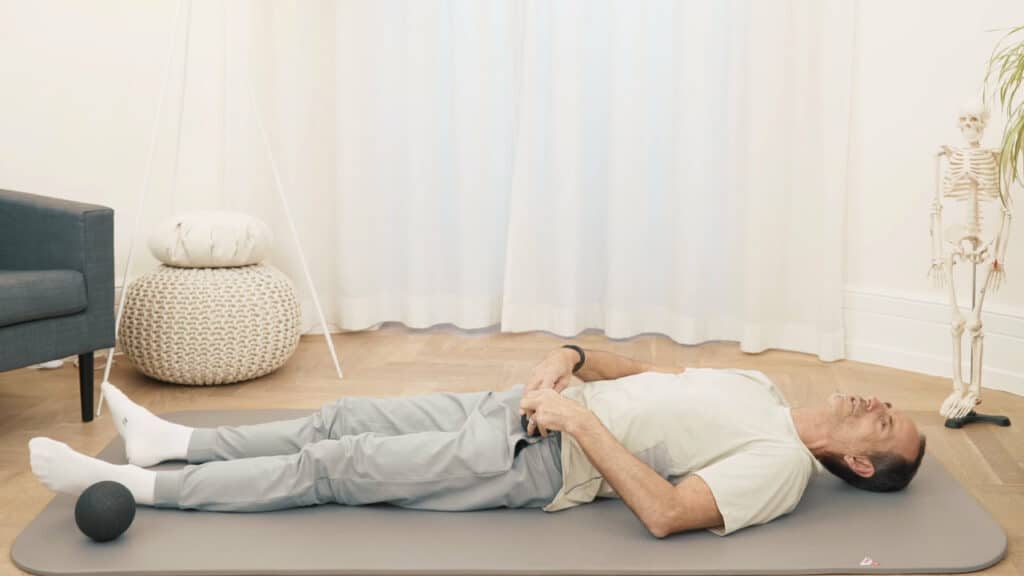 Roland Liebscher-Bracht liegt auf dem Rücken auf einer Matte. Beide Beine sind ausgestreckt und mit beiden Händen hält er die Mini-Kugel an der Leiste, um seinen Hüftbeuger damit zu massieren.