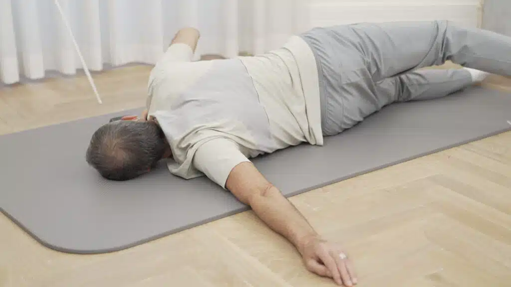 Roland Liebscher-Bracht liegt auf dem Bauch auf einer Matte. Sein linker Arm ist zur Seite ausgestreckt, die Handfläche am Boden. Er dreht den Körper auf die linke Schulter, um den Brustmuskel zu dehnen.