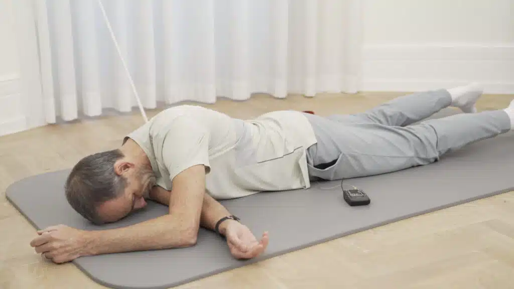 Roland Liebscher-Bracht liegt auf dem Bauch und hat seinen rechten Arm unter seinem Körper nach links rausgelegt. Der linke Arm hilft beim Aufstützen. So dehnt er die Muskulatur rund ums Schulterblatt.