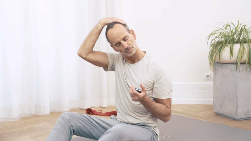 Roland Liebscher-Bracht sitzt auf dem Boden und zieht mit der rechten Hand seinen Kopf zur Seite.