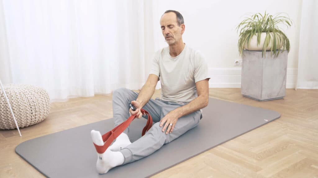 Roland Liebscher-Bracht sitzt auf dem Boden. Sein linkes Bein ist ausgestreckt und er zieht mit einer roten Schlaufe seinen Fuß zurück.