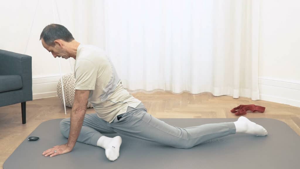 Roland Liebscher-Bracht hat am Boden eine Übungsposition eingenommen. Sein linkes Bein ist nach hinten ausgestreckt und das rechte Bein liegt angewinkelt vor ihm, während er seinen Oberkörper mit beiden Armen abstützt.