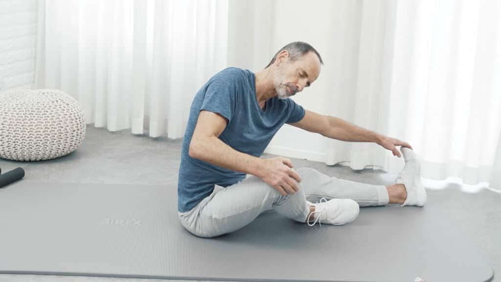Roland Liebscher-Bracht sitzt auf der Matte. Sein rechtes Bein hat er lang vor sich ausgestreckt, seinen linken Fuß an dessen Innenseite angelegt. Er beugt sich nach vorne, um mit der rechten Hand auch den Fuß des langgestreckten Beines packen zu können.