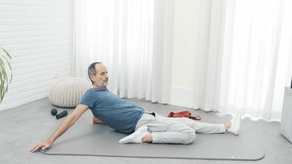 Roland Liebscher-Bracht sitzt auf der Matte. Sein linkes Bein hat er bis zu seinem Gesäß angewinkelt, sein rechtes lang vor sich ausgestreckt. Mit dem Oberkörper lehnt er sich weiter zurück.