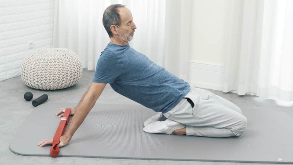 Roland Liebscher-Bracht sitzt auf seinen Fersen und geht mit dem Oberkörper immer weiter nach hinten. Er hält hinter seinem Rücken die Hände in der Übungs-Schlaufe, die Knie bleiben auf dem Boden.