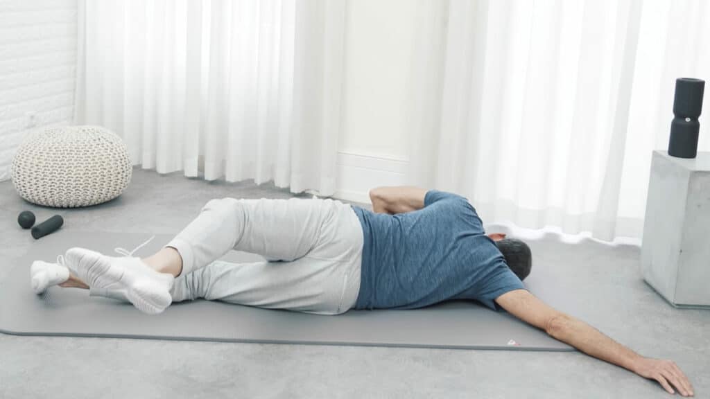 Roland Liebscher-Bracht liegt auf der rechten Körperseite. Sein rechter Arm liegt langgestreckt nach hinten raus. Sein linkes Bein hält er angehoben leicht nach hinten.