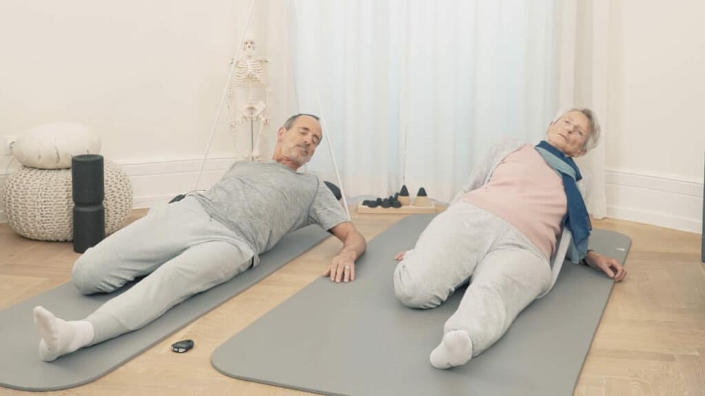 Schmerzspezialist Roland Liebscher-Bracht und seine Mutter Ruth sitzen jeweils auf ihrer grauen Übungsmatte: Ihr linkes Bein ist jeweils nach vorne ausgestreckt, währen sie ihre rechtes zu ihrem Gesäß hoch anwinkeln. Die Oberkörper lehnen sie beide nach hinten.